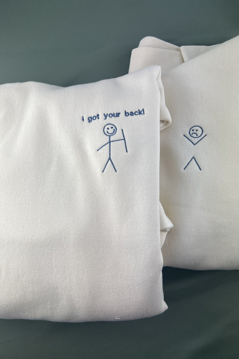 Two ivory hoodies side by side with the embroidered I got your back smiling stick figure and a matching sad stick figure