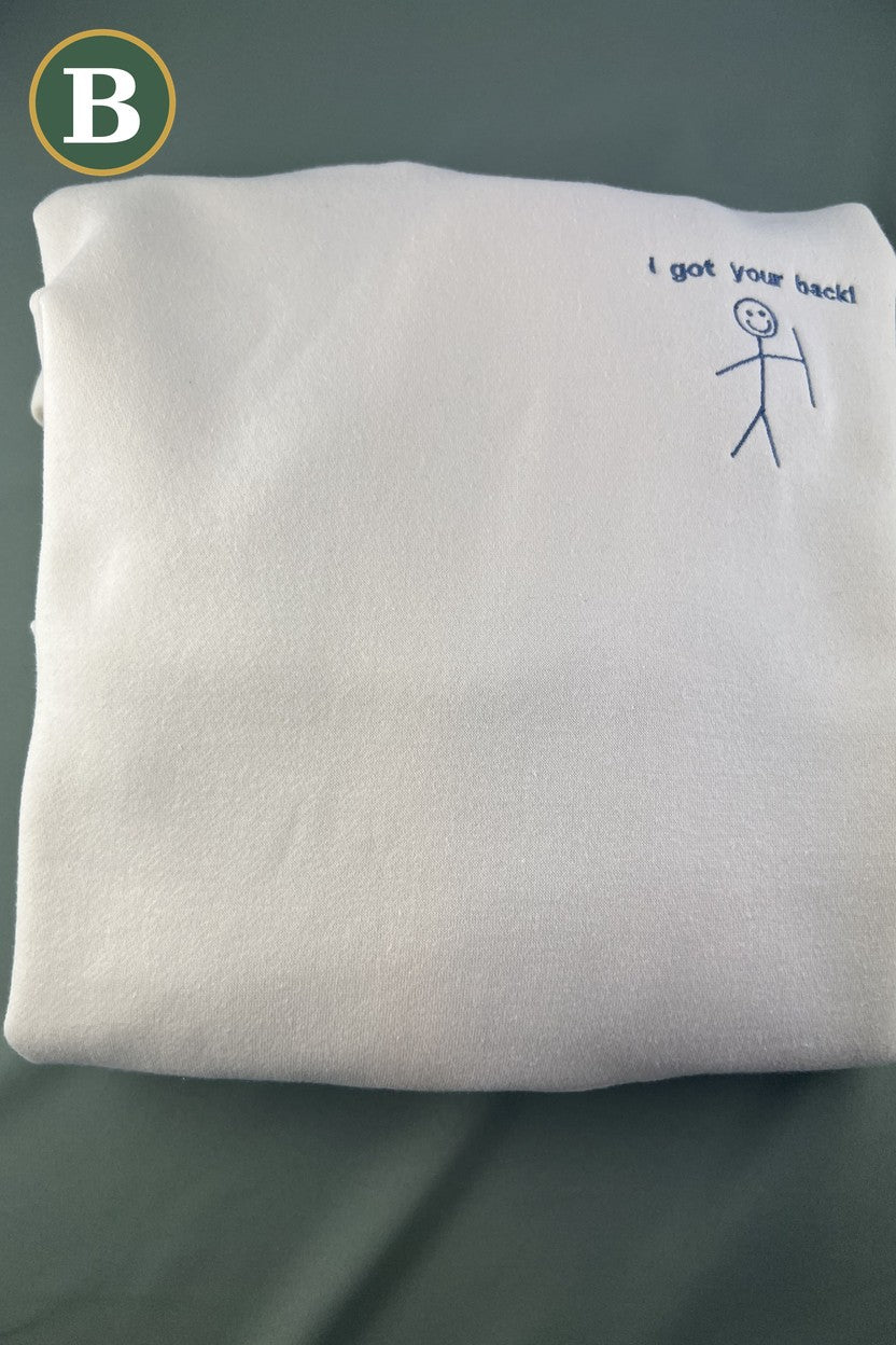Folded ivory hoodie with the embroidered I got your back smiling stick figure on the chest, option B