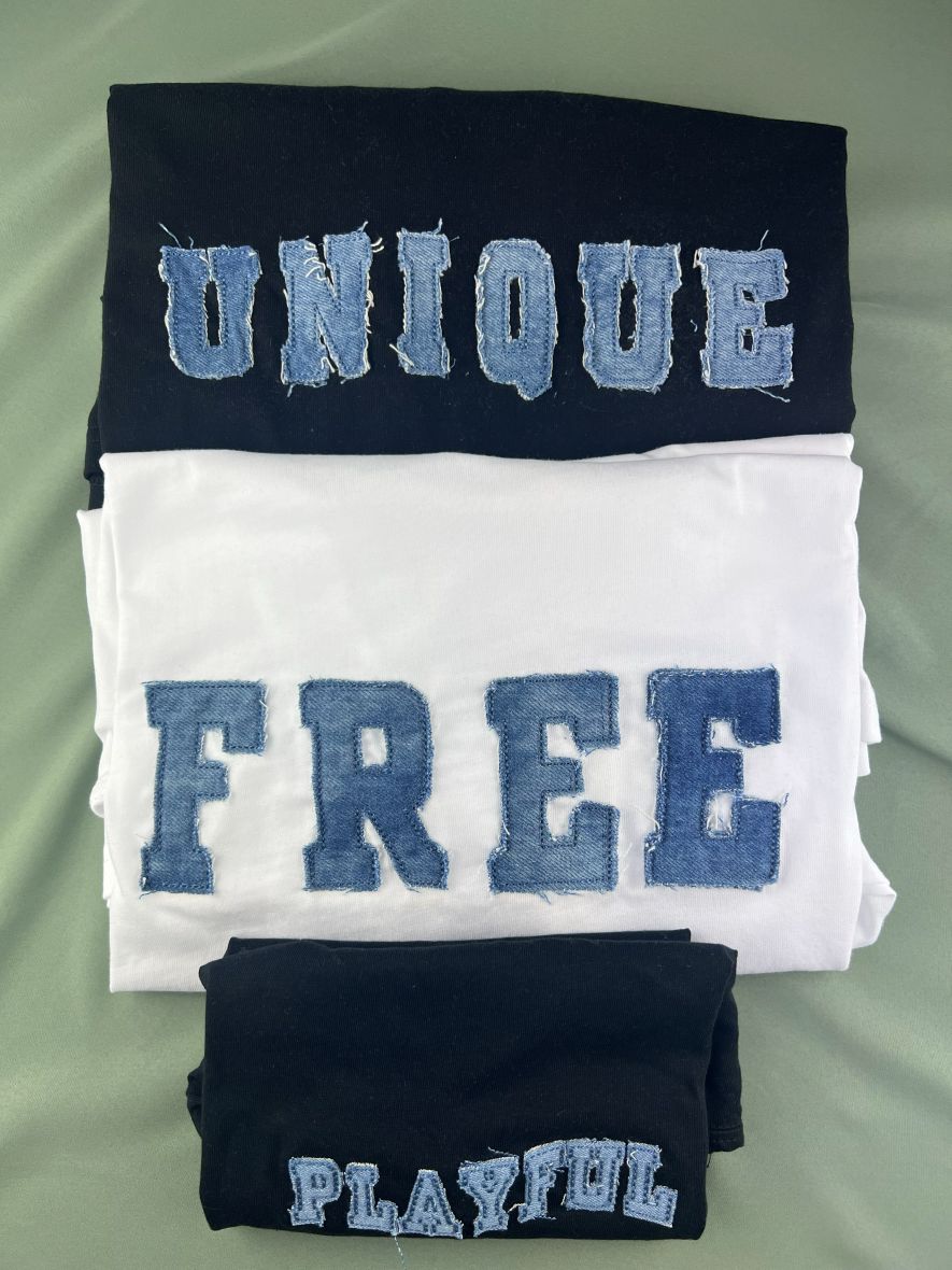 Flat-lay of black and white tees with denim-patch lettering reading UNIQUE, FREE and PLAYFUL